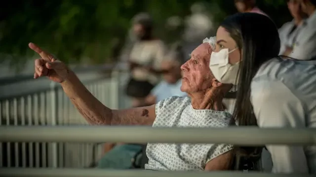 Foto de um senhor sentado apontado para a frente enquanto uma mulher de máscara e vestida de branco olha para onde ele aponta.