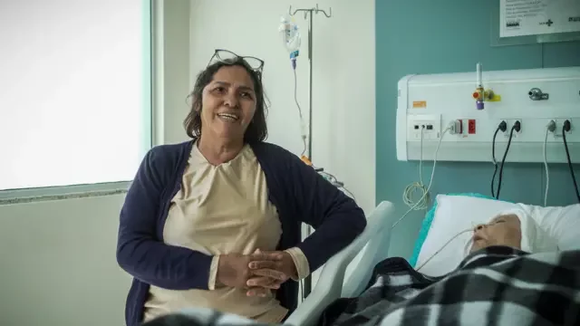 Foto de uma senhora de pé, ao lado de uma cama de hospital onde está deitado um senhor.